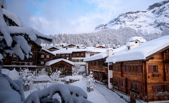 Wooden homes with snow on top of them, sitting in the valley of a snowy mountain